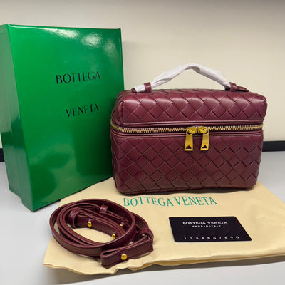 Maroon woven handbag with gold zippers, matching belt, and green Bot tega Ven eta box on a beige surface.