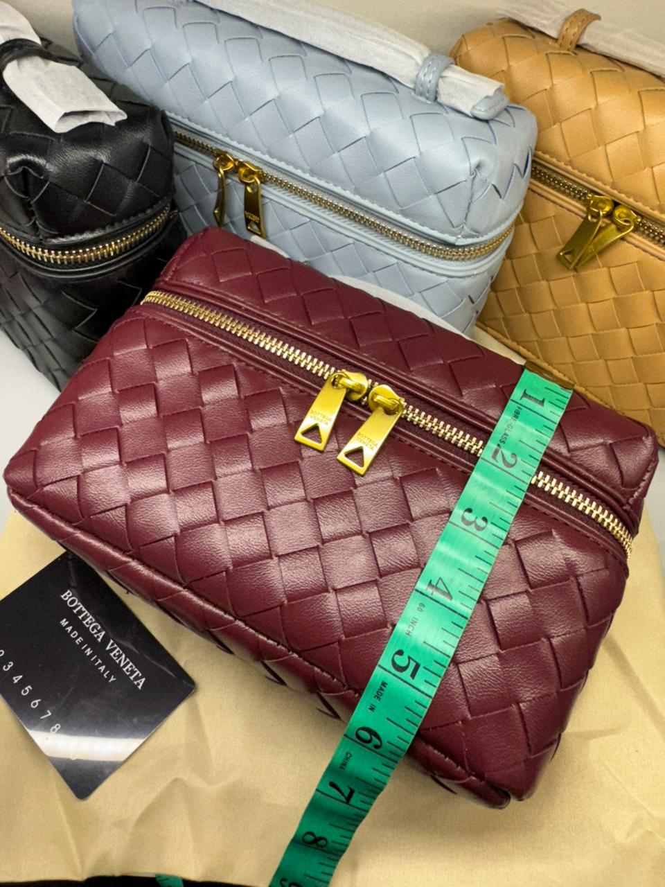 Maroon woven handbag with gold zipper and green measuring tape on a beige surface, with other bags in the background.