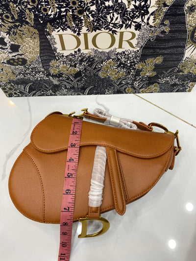 Brown saddle bag with a pink measuring tape draped over it, against a D ior branded background.