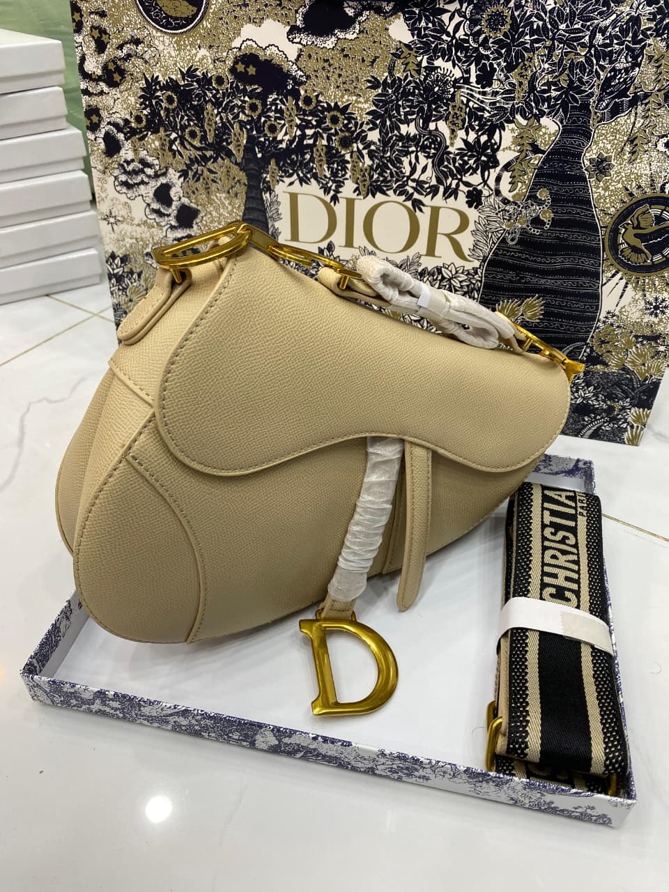 Beige saddle-shaped handbag with gold accents on a Di or branded stand.