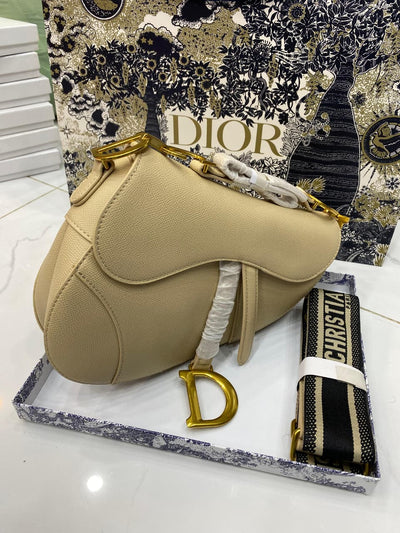 Beige saddle-shaped handbag with gold accents on a Di or branded stand.