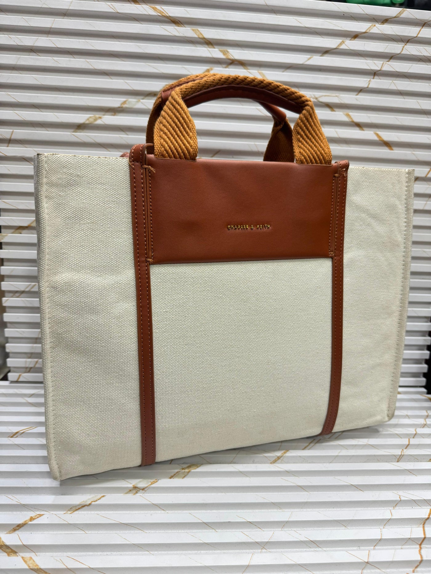 Beige tote bag with brown leather accents on a corrugated metal background
