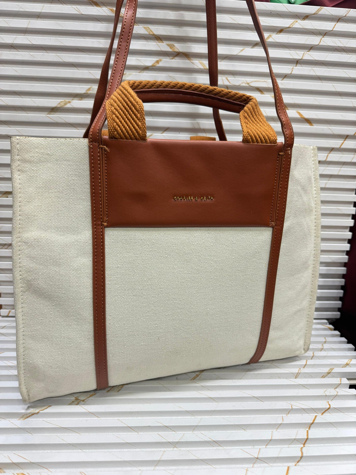 Beige tote bag with brown leather accents on a corrugated metal background