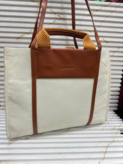 Beige tote bag with brown leather accents on a corrugated metal background