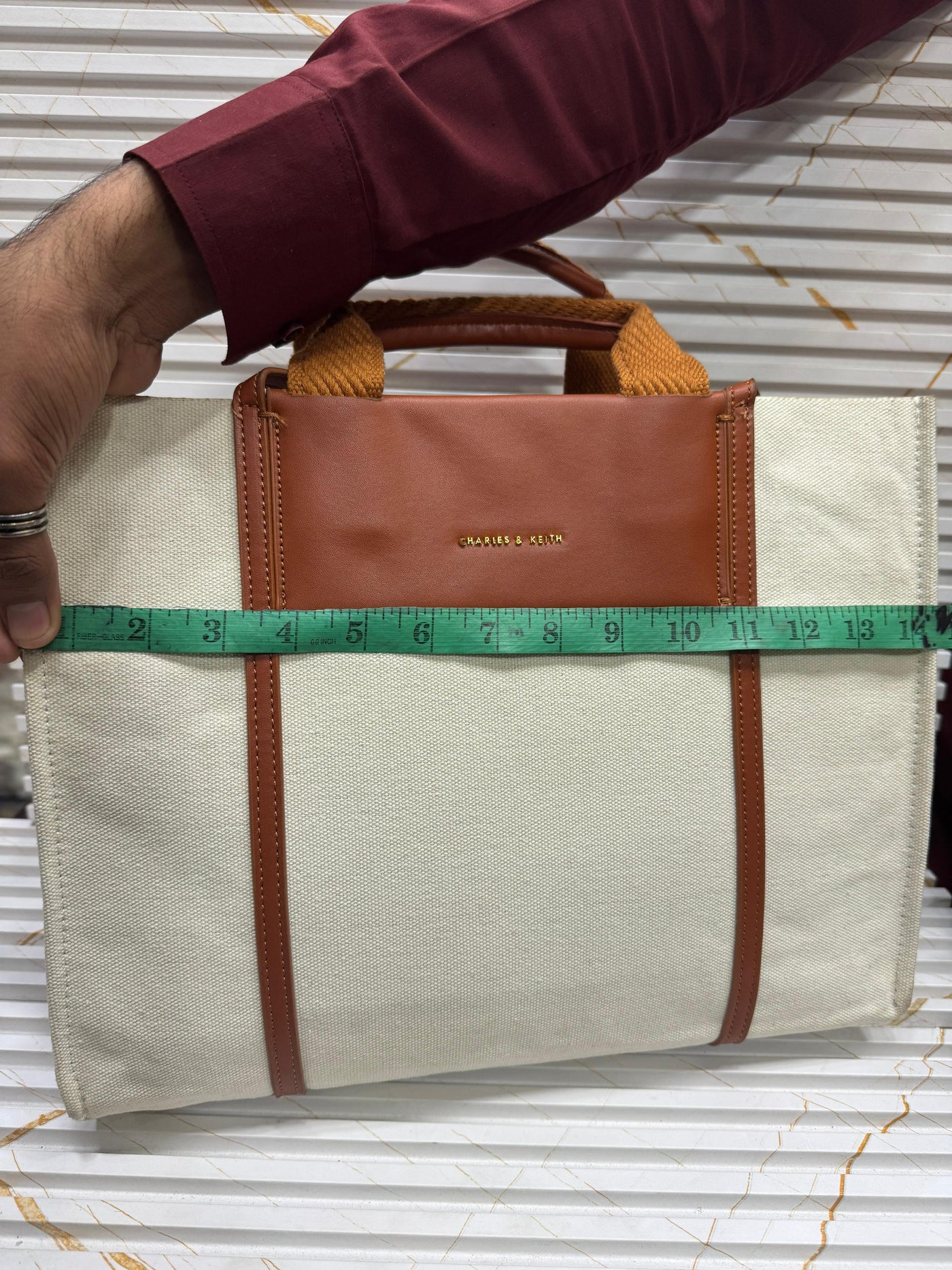 Beige bag with brown accents being measured with a green tape measure on a corrugated metal background.