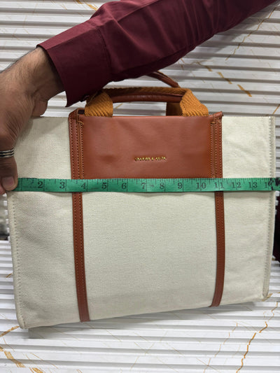 Beige bag with brown accents being measured with a green tape measure on a corrugated metal background.