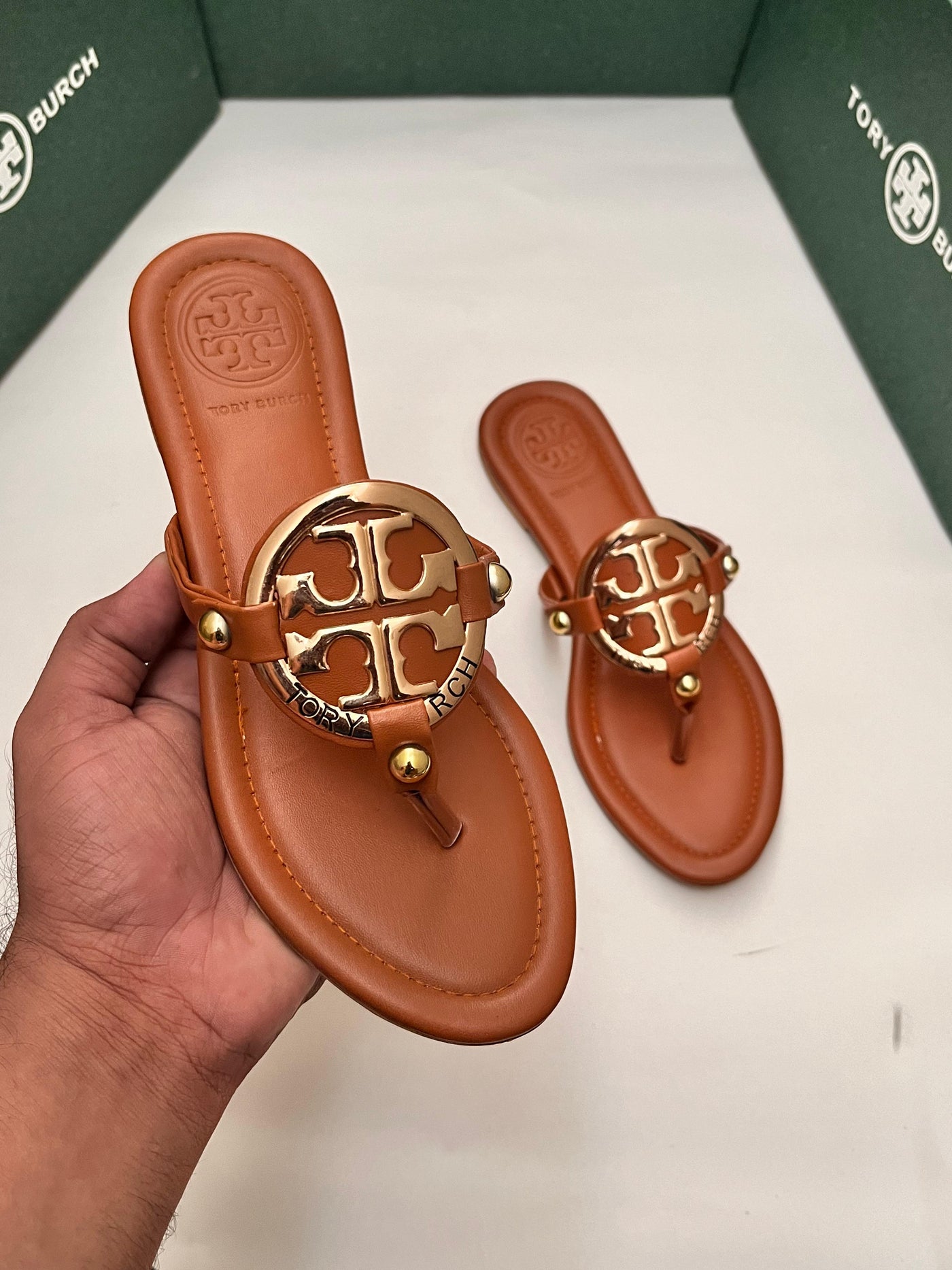 Brown sandal with gold logo held by a hand, brand box in the background