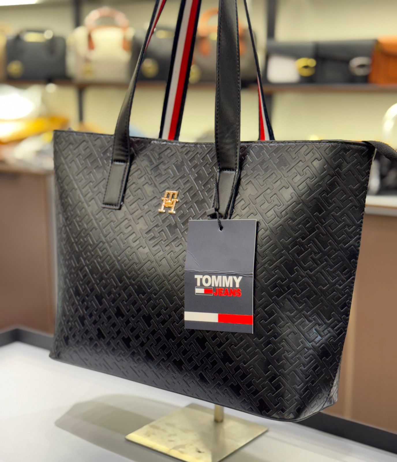 Black textured tote bag with Tom my Hil figer tag on a stand in a store setting