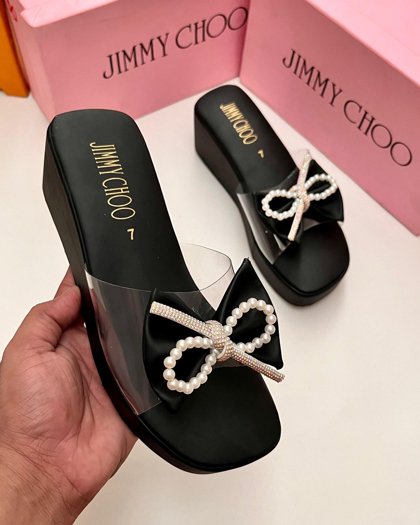 Black sandals with pearl bows held by a hand, with Jim my Ch oo packaging in the background.